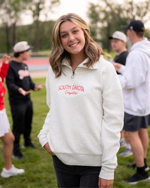 Young woman wearing a white quilted quarter zip with red 'South Dakota Coyotes' embroidered on chest, standing outdoors with blurred people in the background.