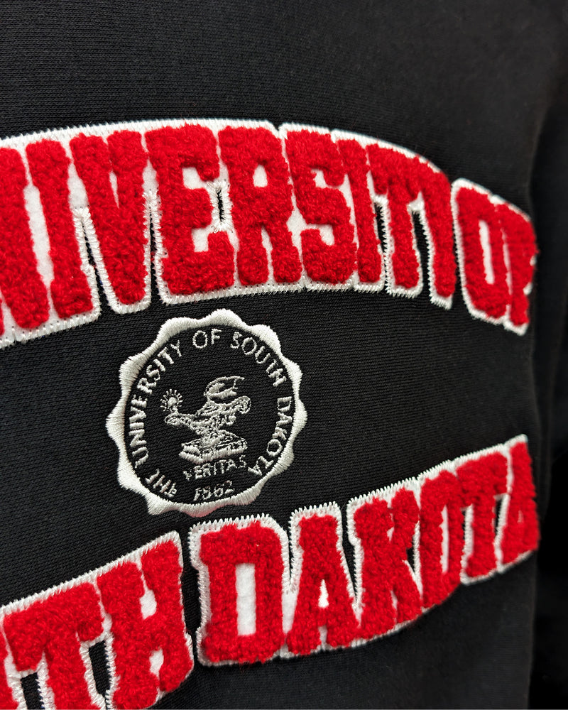 Close-up of black crew with chenille red and white letters, 'THE UNIVERSITY OF SOUTH DAKOTA,' with white embroidered university seal in the center. 