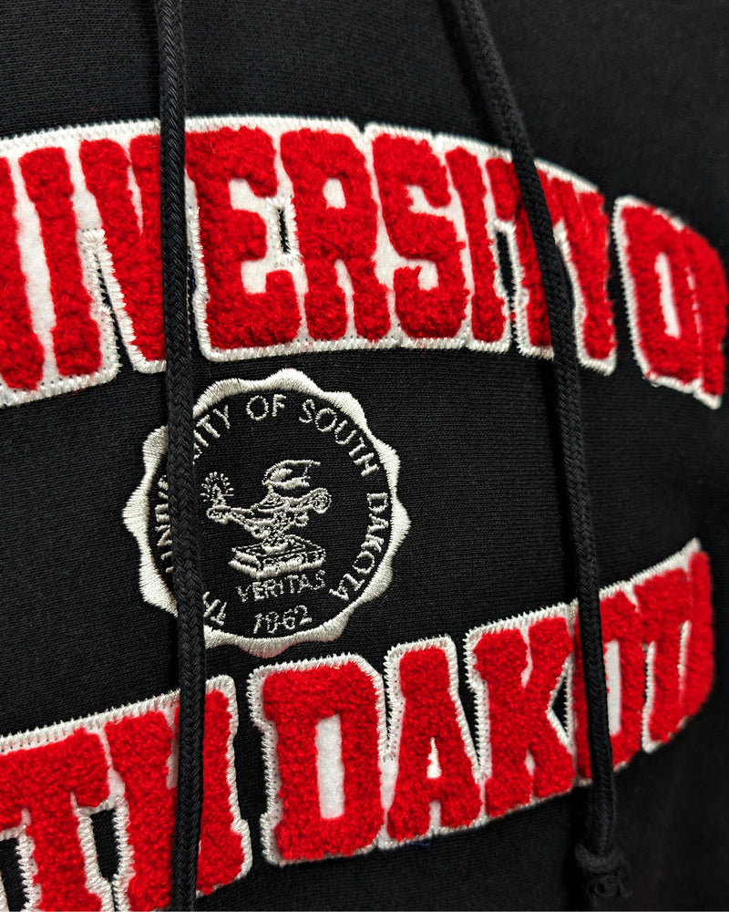 Close-up of black hoodie with chenille red and white letters, 'THE UNIVERSITY OF SOUTH DAKOTA,' with white embroidered university seal in the center. 