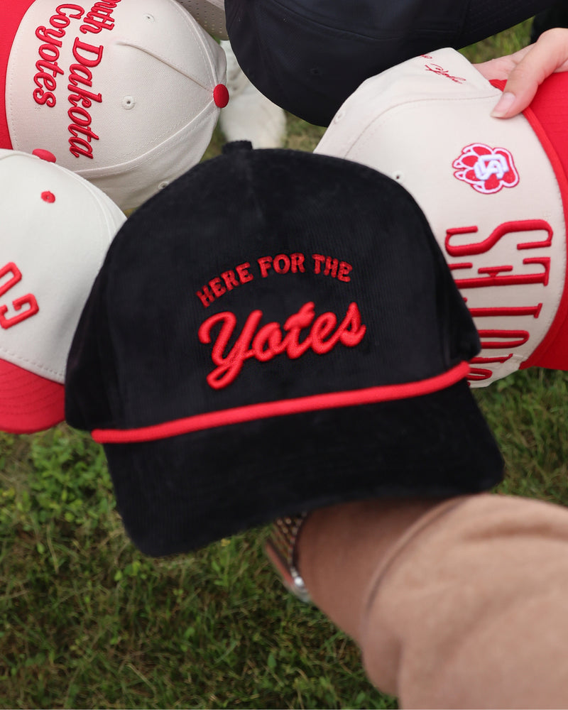 Black corduory cap with red rope and red embroidered text 'HERE FOR THE Yotes' held by a person on grass.