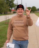 Person wearing a brown sweatshirt with 'UNIVERSITY OF SOUTH DAKOTA USD COYOTES' embroidered on it, making a coyotes sign outdoors.
