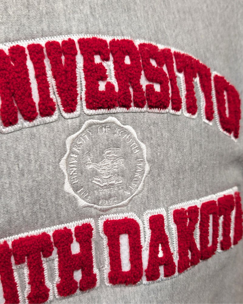 Close-up of gray crew with chenille red and white letters, 'THE UNIVERSITY OF SOUTH DAKOTA,' with white embroidered university seal in the center. 