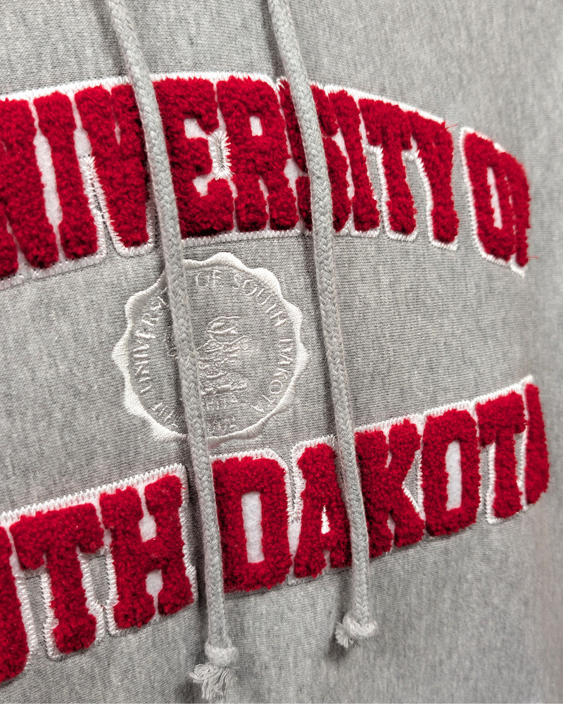 Close-up of gray hoodie with chenille red and white letters, 'THE UNIVERSITY OF SOUTH DAKOTA,' with white embroidered university seal in the center. 
