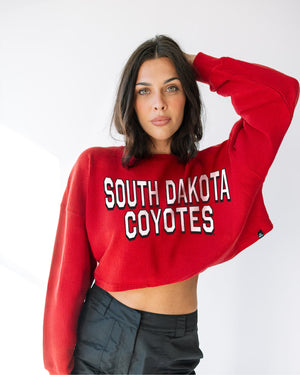 Person wearing a red cropped corded crew with white 'South Dakota Coyotes' across chest.