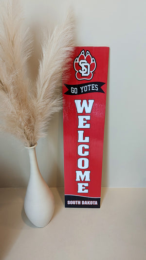 Red vertical porch leaner sign with SD Paw logo above text that says 'GO YOTES! WELCOME' and 'SOUTH DAKOTA' next to a large tan vase with pampas grass