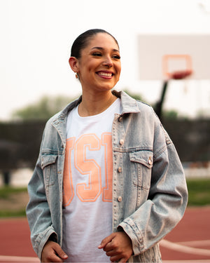 Woman standing on an outdoor basketball court wearing the white cuffed tee with coral USD letter on front and a jean jacket over it