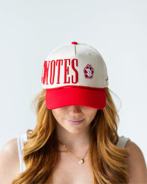 Front of woman wearing a cream colored hat with red bill and rope and large embroidered serif text 'COYOTES' with the SD Paw logo. Text starts on side of head rather than only at front.
