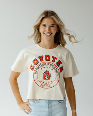 Woman wearing an oatmeal t-shirt with 'Coyotes' and 'University of South Dakota' print in red in a circular design.