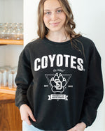 Woman wearing the black corded crew with white text and design, 'COYOTES Go Yotes! Est. 1862 UNIVERSITY OF SOUTH DAKOTA,' and the SD Paw logo with a triangle design behind.