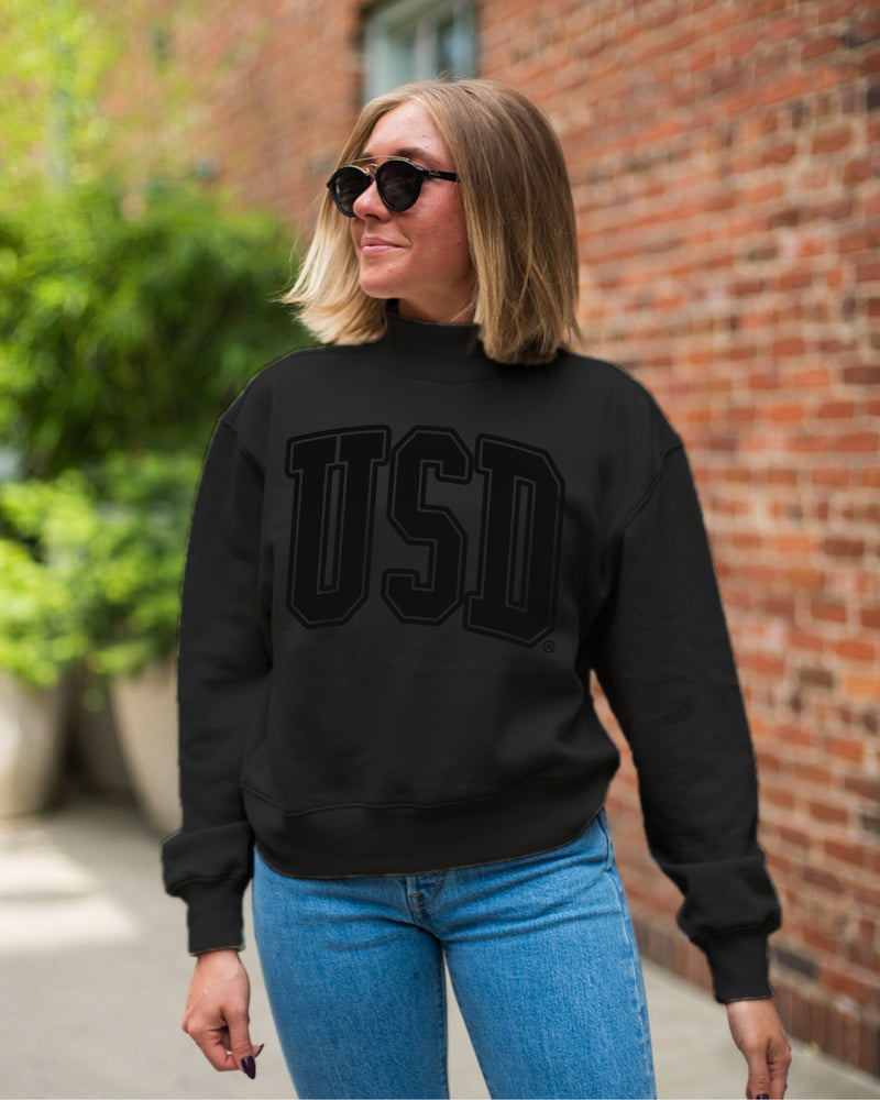 Woman wearing black mock neck cropped crew with large black, 'USD,' across chest.