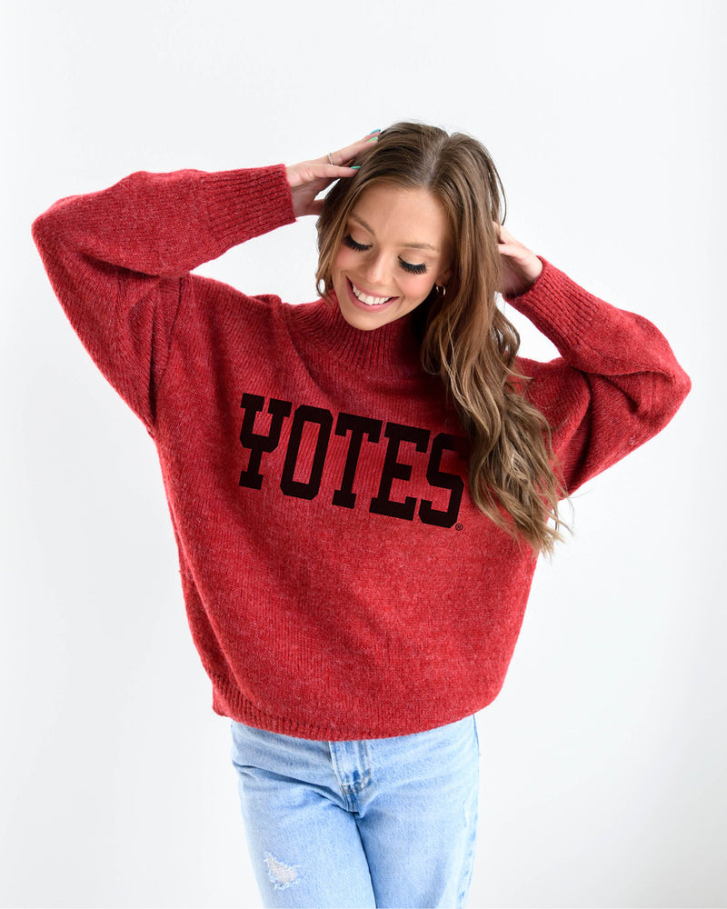 Woman wearing jeans and red turtleneck sweater with black text, ' YOTES,' across chest. 