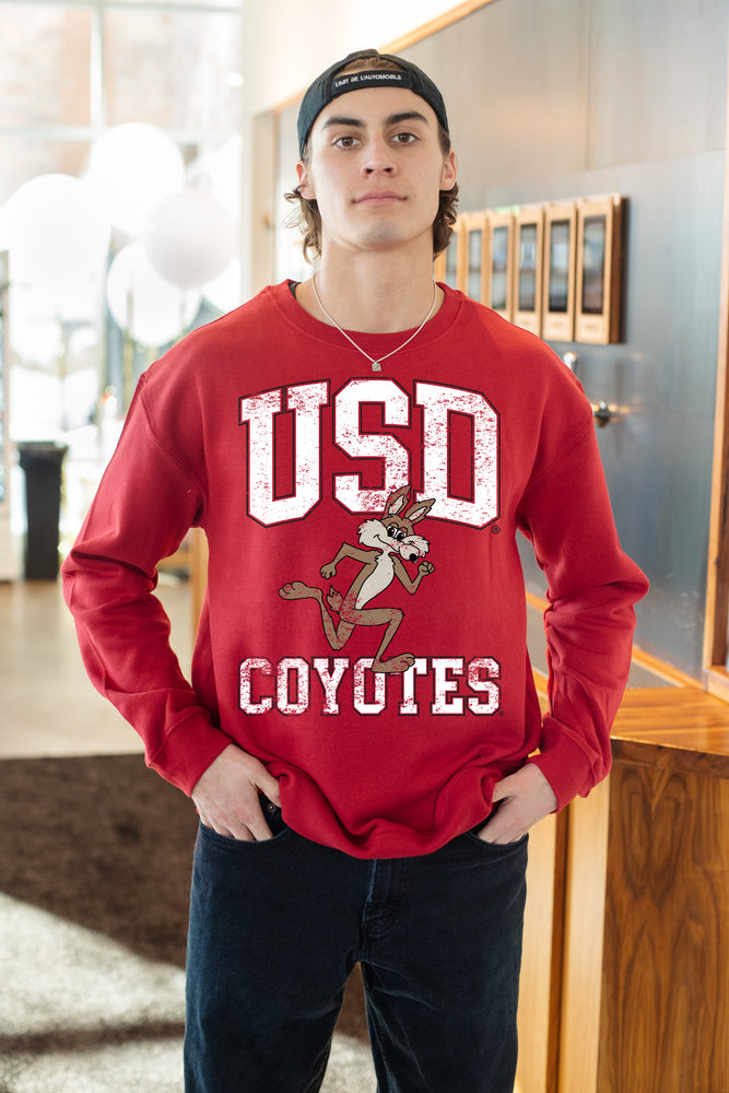 
                
                    Load image into Gallery viewer, Man standing with hands in his pockets wearing the Unisex Red Vintage Charlie crew that has white text that says, &amp;#39;USD COYOTES&amp;#39; with vintage Charlie running between USD and Coyotes
                
            