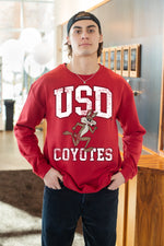 Man standing with hands in his pockets wearing the Unisex Red Vintage Charlie crew that has white text that says, 'USD COYOTES' with vintage Charlie running between USD and Coyotes